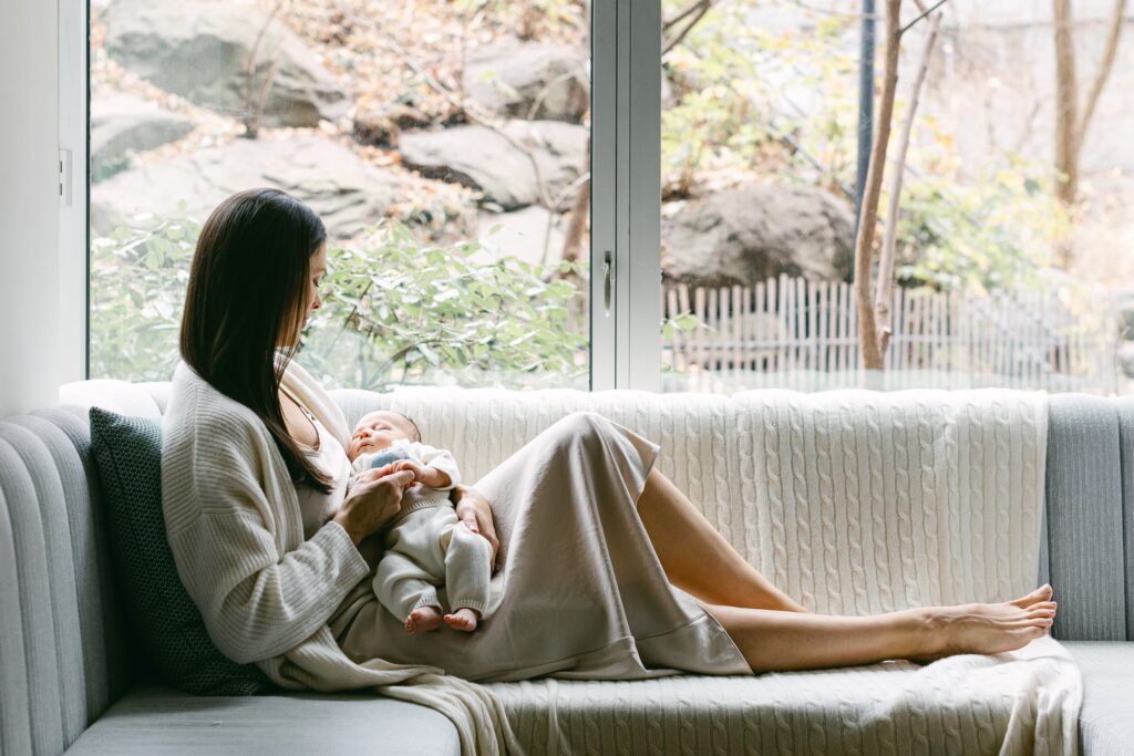 Mom in neutral sweater in dress, resting on window seat with legs stretched out, and newborn baby in lap