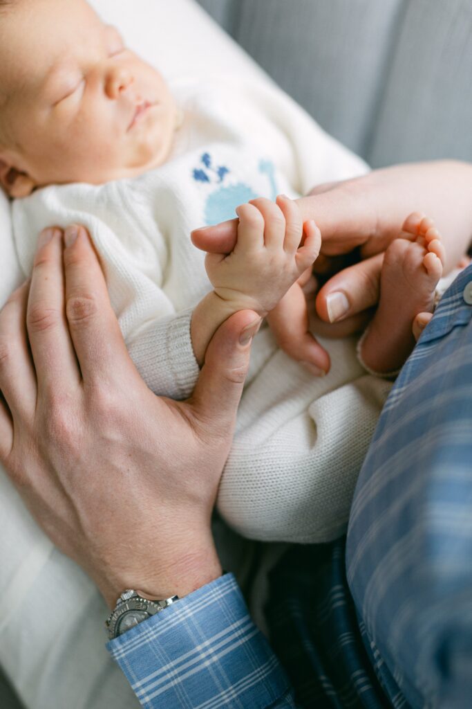 Close-up shot of newborn baby in white knits holding his father's finger.