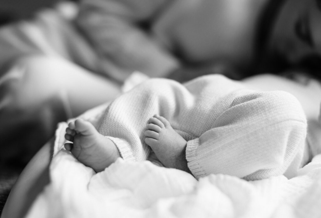 Black and white image of newborn baby's legs and feet, with mom resting near him in background.