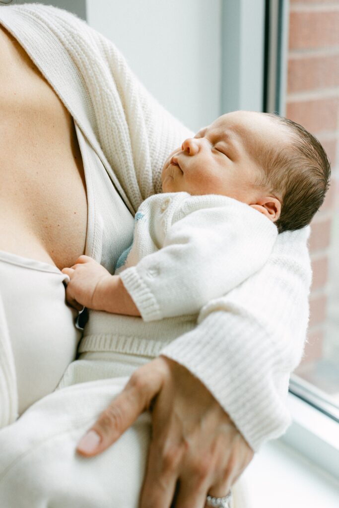 Newborn baby in a white knit sweater set holds onto his mother's dress.