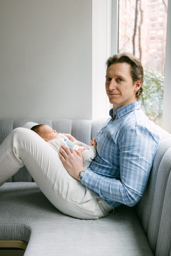 Dad sitting on couch near window with newborn baby in lap.