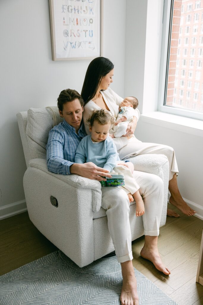New York family of four with toddler and newborn baby, cuddled together in a white rocking armchair