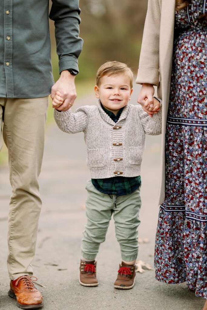 Close-up shot of smiling toddler boy holding both his mom and dad's hand, each parent lining the edge of the photo. 