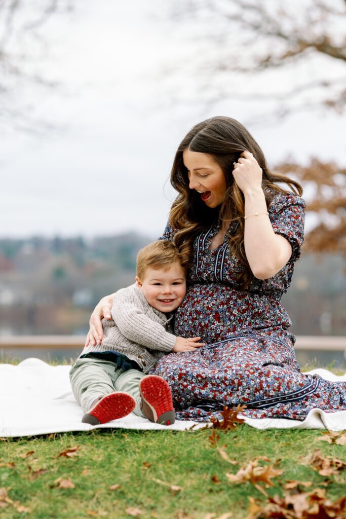 Brunette mom sitting on blanket in the park with toddler boy who is smiling and wrapping his arms around her belly. 