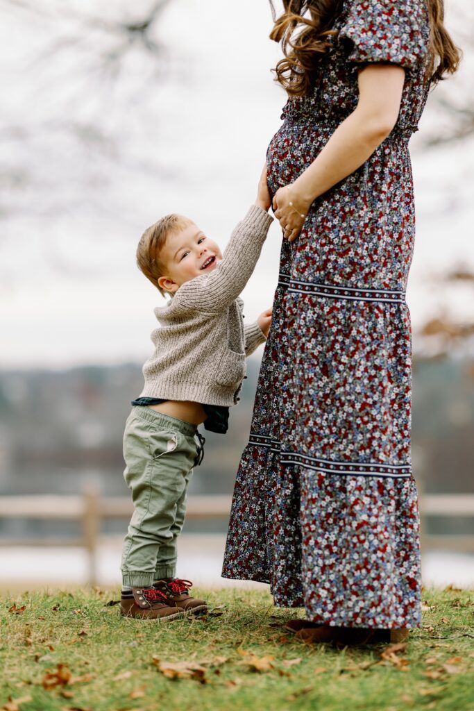 Mom in long floral dress with toddler boy resting hand on her pregnant belly and smiling at the camera.