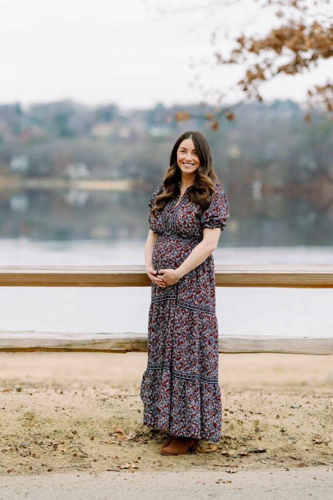 Brunette Mom standing by railing in long floral dress smiling at camera and holding onto pregnant belly. 