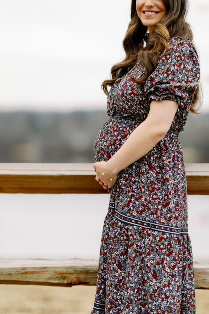 Brunette mom in long floral dress clutching belly and smiling at the camera.