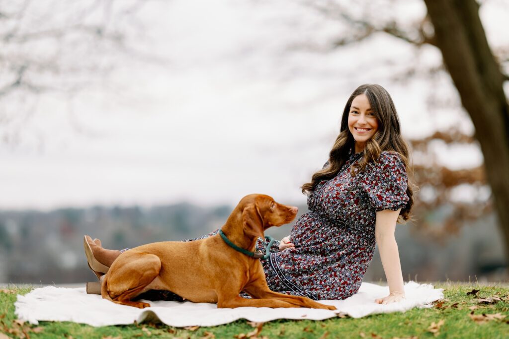 Brunette smiling Mom sitting on blanket in the park with brown Vizsla dog. 