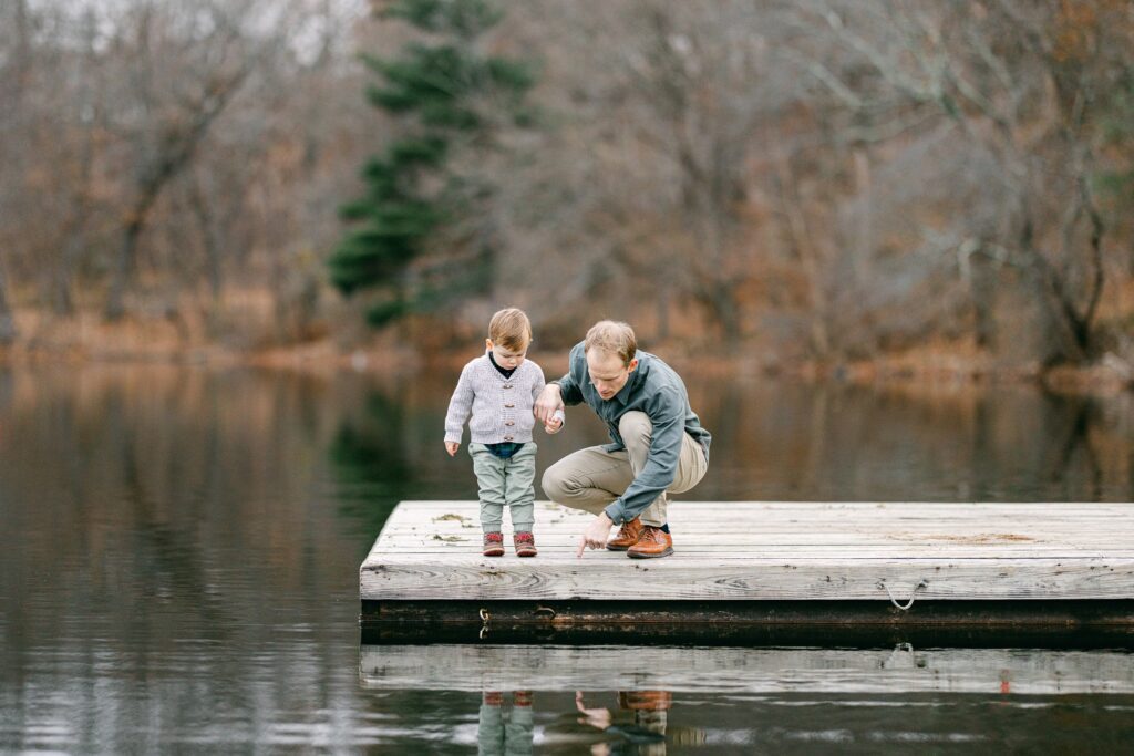 Dad and toddler boy standing on dock and looking at something in the water. 