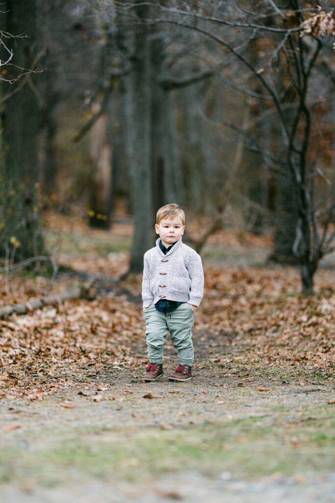 Toddler boy standing in the woods, hands in pockets, staring in the camera. 