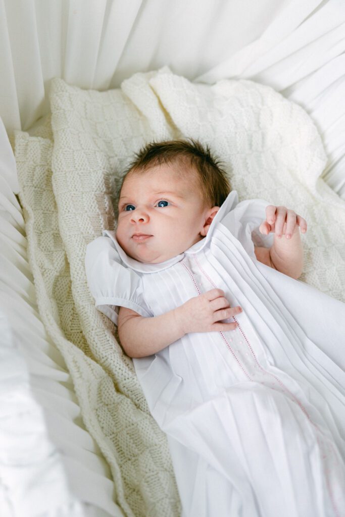 Baby with brown hair laying in bassinet wearing white gown and laying on a white blanket.
