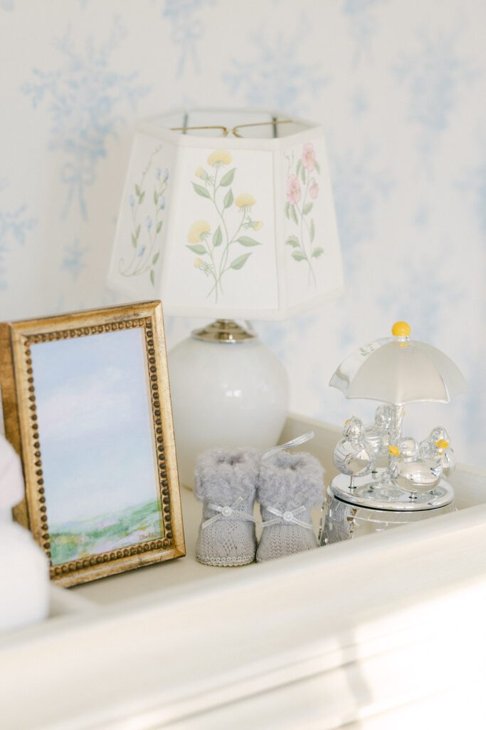 Nursery decor including a gold picture frame, baby booties, silver music player and lamp with florals on lampshade.