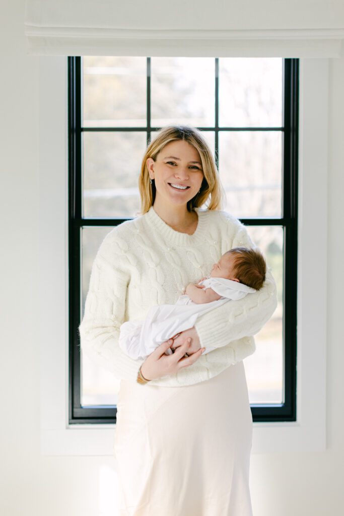 Mom smiling big in all white while holding baby in front of the window. 