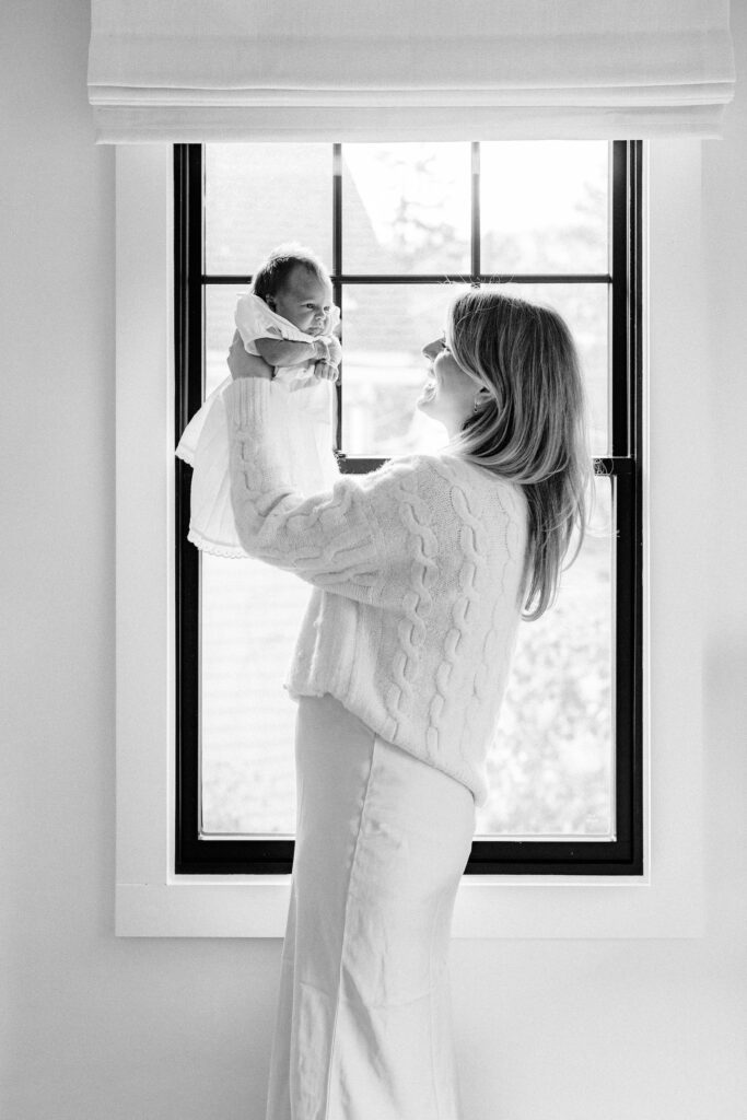 Mom standing in front of window in white sweater and silk skirt, holding baby up at eye level and smiling at her. 