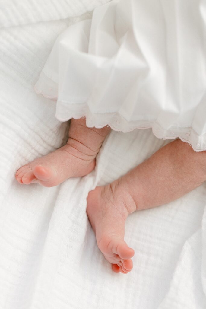 Baby's legs and feet sticking out of white gown while laying on white blanket. 