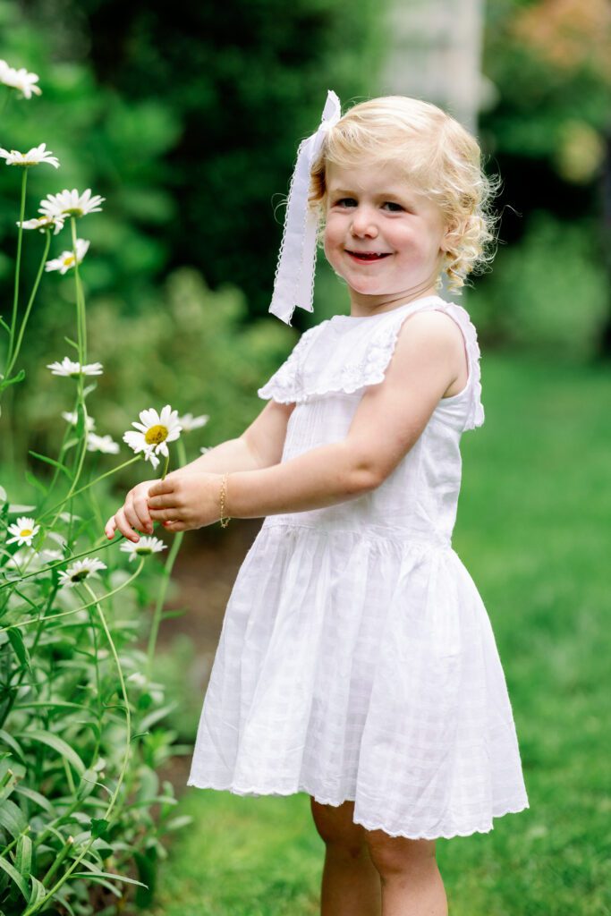 toddler in a white dress surrounded by greenery, holding a flower