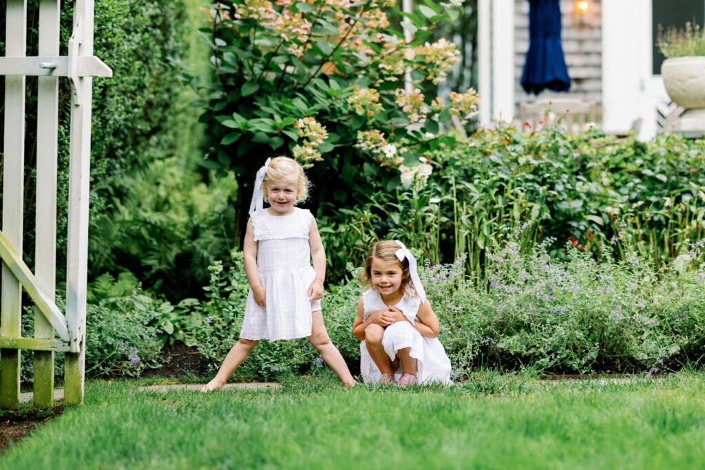 Two young sisters in white dresses surrounded by greenery in their fenced in backyard.