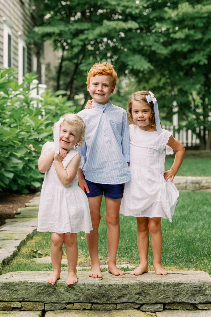 Three siblings stand together in their backyard, dressed in white and blue.