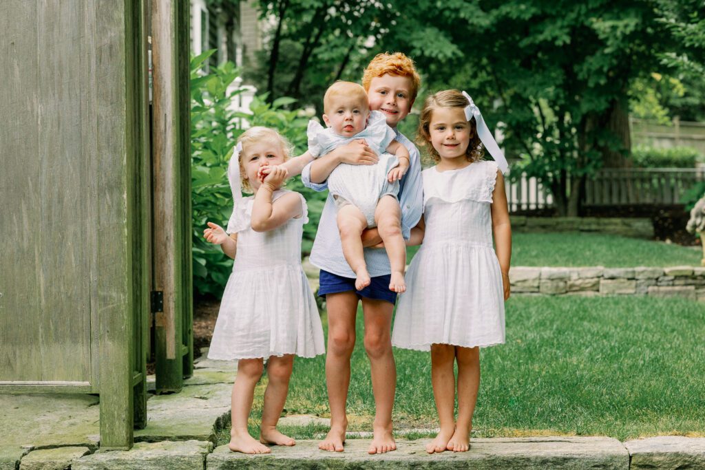 Four siblings in white and blue stand on a step in their backyard, the oldest child holding the youngest, a baby.