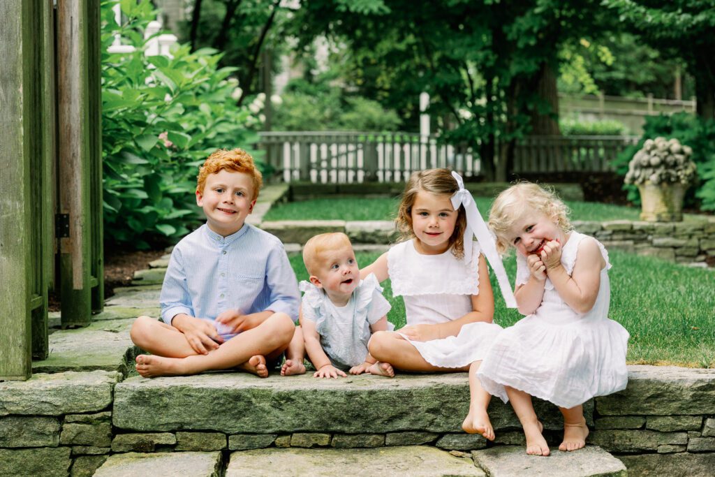 Four siblings in white and blue sit on a step in their backyard, smiling at the camera.