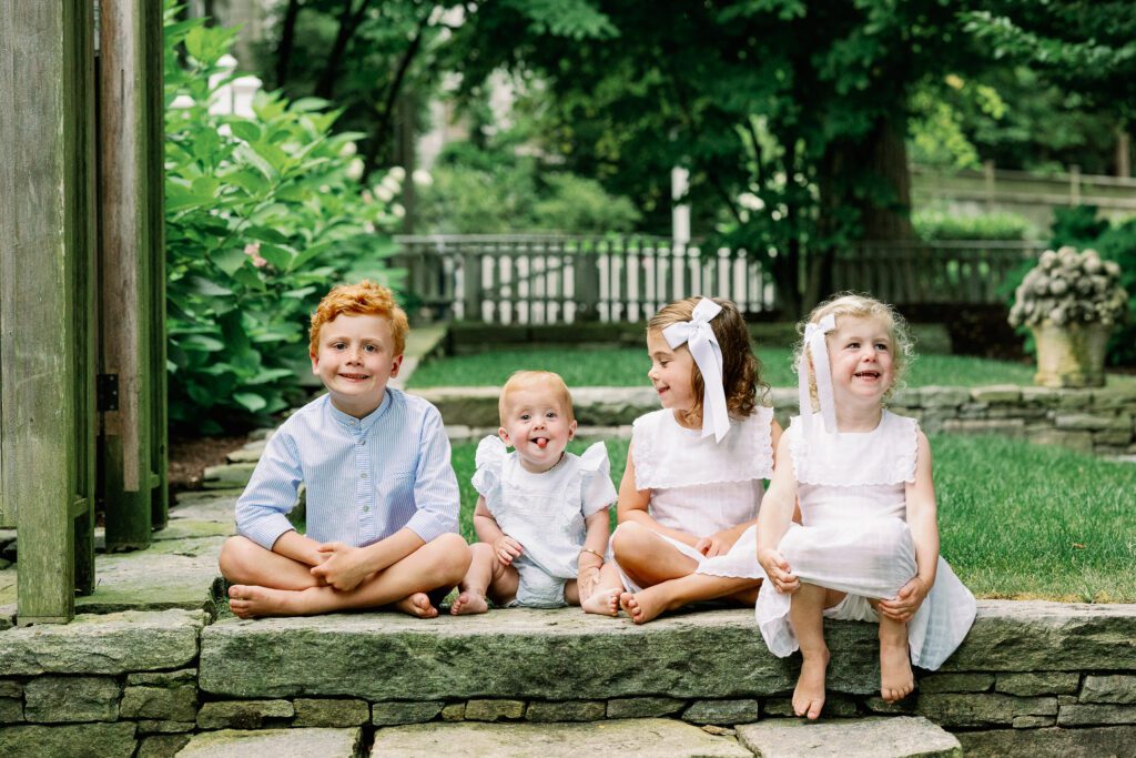 Four siblings in white and blue sit on a step in their backyard, smiling at the camera and each other.