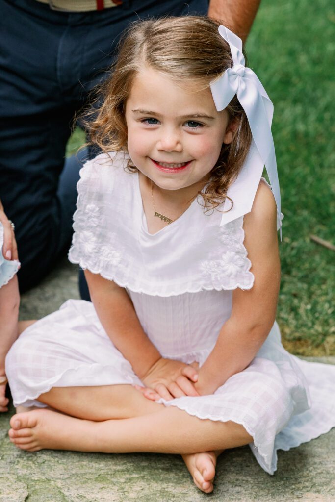 Young girl in a white dress and white bow smiles up at the camera.