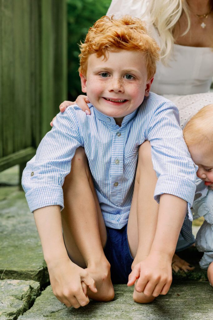 Young red-headed boy with freckles sits holding his feet, dressed in blue.