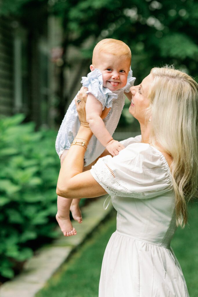 Blond mom in a white dress lifts her baby in blue up to eye level, while baby looks at camera with her tongue out.