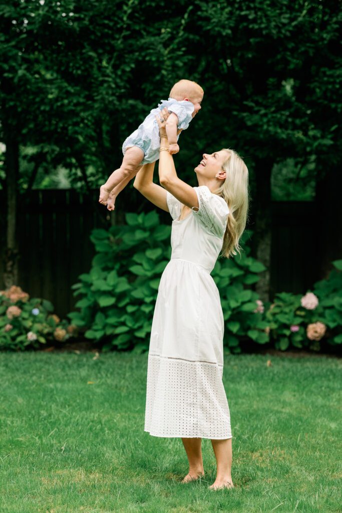 Blond mom in a white dress lifts her baby in blue over her head in the backyard.