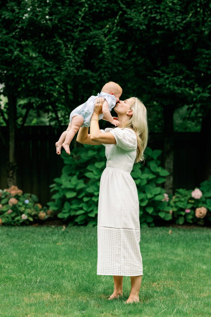 Blond mom in a white dress lifts her baby in blue up in the air and gives her a kiss, among the greenery in the backyard.