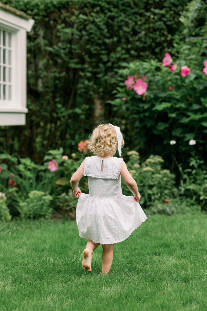 Toddler in white dress runs away from camera holding her dress as she dances through her backyard.