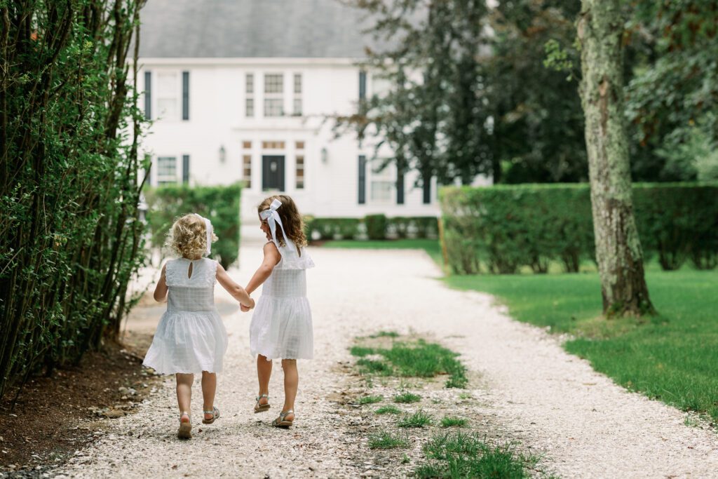 Wide shot of two sisters in white dresses holding hands and running down the shelled driveway of their Nantucket vacation home.