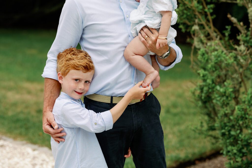 Red-headed boy holds his baby sister's foot, while dad wraps one arm around each child.