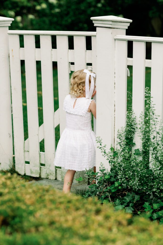 Toddler in a white dress enters a garden through a white fence, peeking through the fence to see what lies beyond.