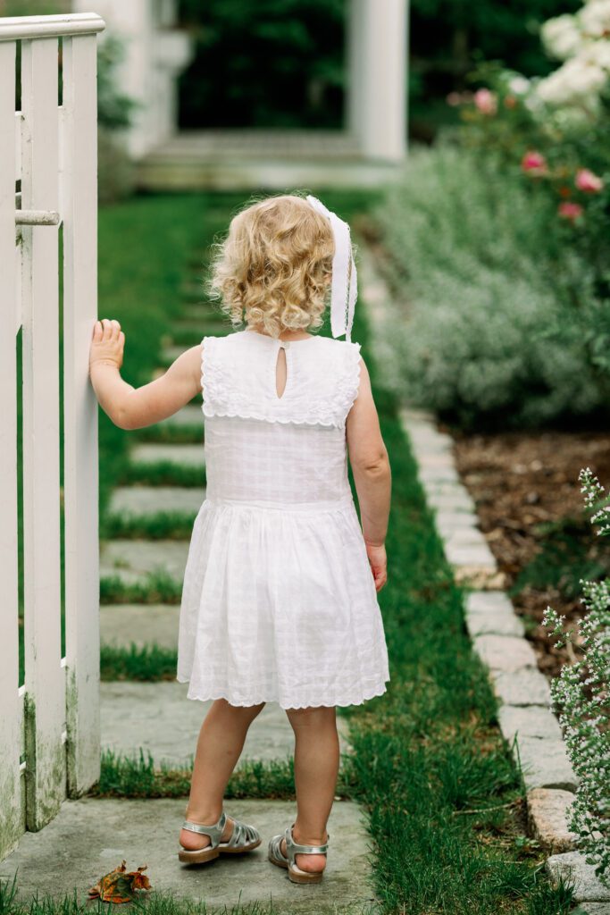 Toddler in a white dress enters a garden through a white fence.