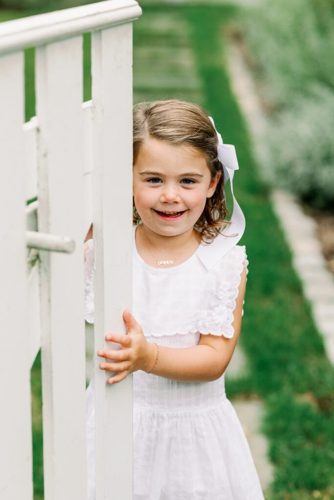 Young girl in a white dress smiles at camera after entering a garden with a white fence.