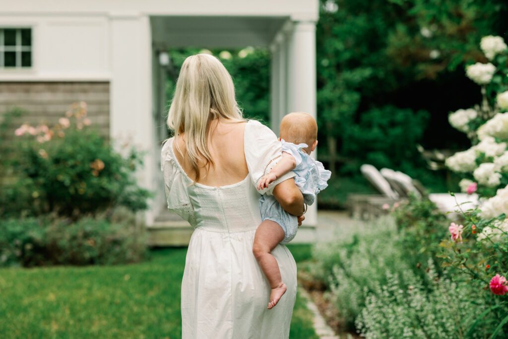 Mom in white dress with blond hair walks away from camera into a garden, holding baby girl with strawberry blond hair.