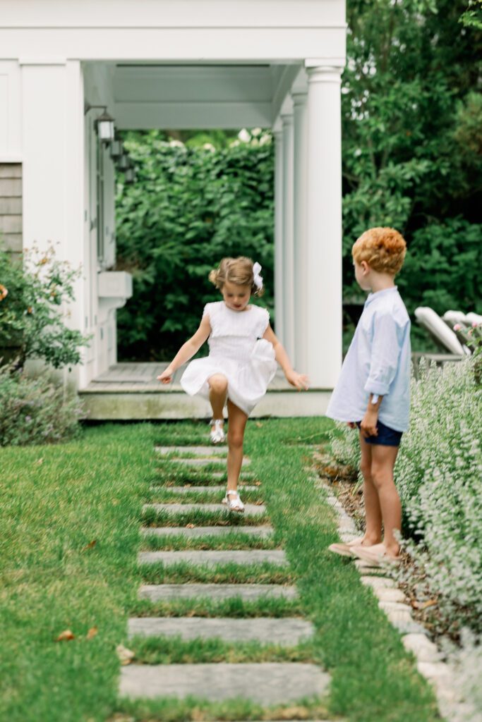 Blurred image showing a young girl in white dress and bow skipping along the step stones in her backyard, as her red-headed brother watches.
