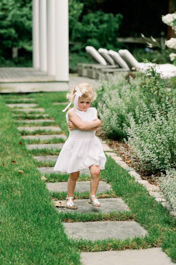 Toddler in a white dress crosses her arms and runs down the step stones in her backyard, with a fussy expression on her face.