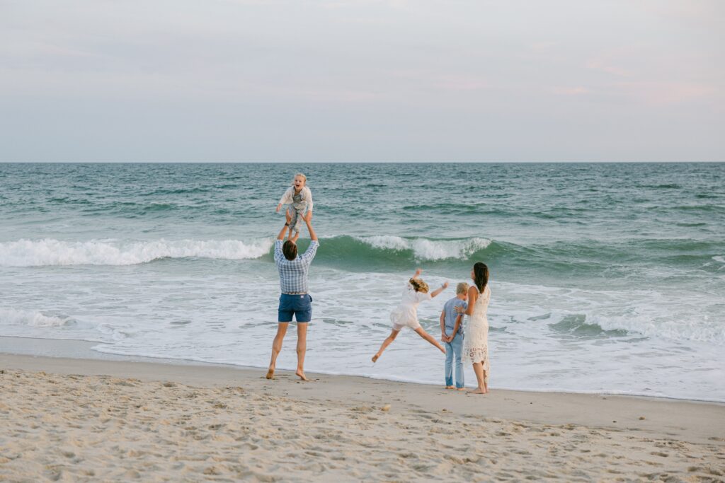 Distance shot of family standing in front of the ocean, with dad lifting the youngest blonde daughter into the air. 
