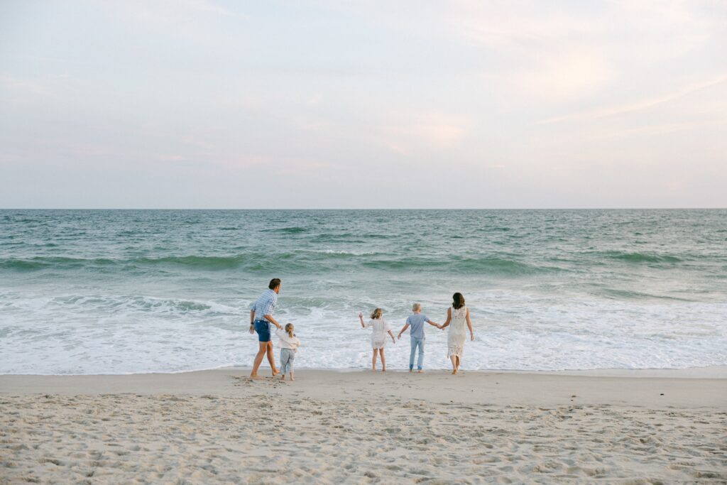 Distance shot of family standing in front of the ocean, with mom and oldest two children holding hands and standing in a row. 
