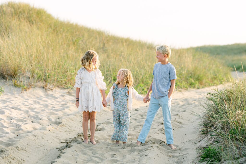 Three blond kids standing in a row holding hands in the sand dunes, all looking over at one another. 