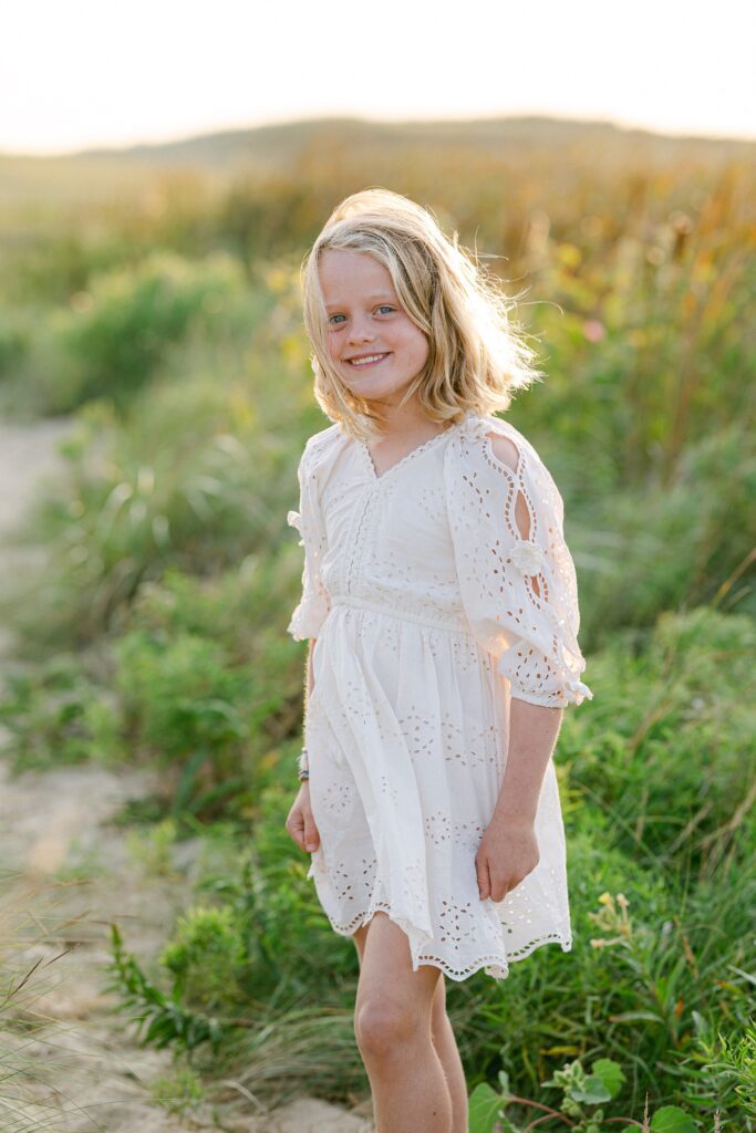 Close-up of middle blonde daughter standing in the sand dunes smiling at the camera with wind-blown hair. 