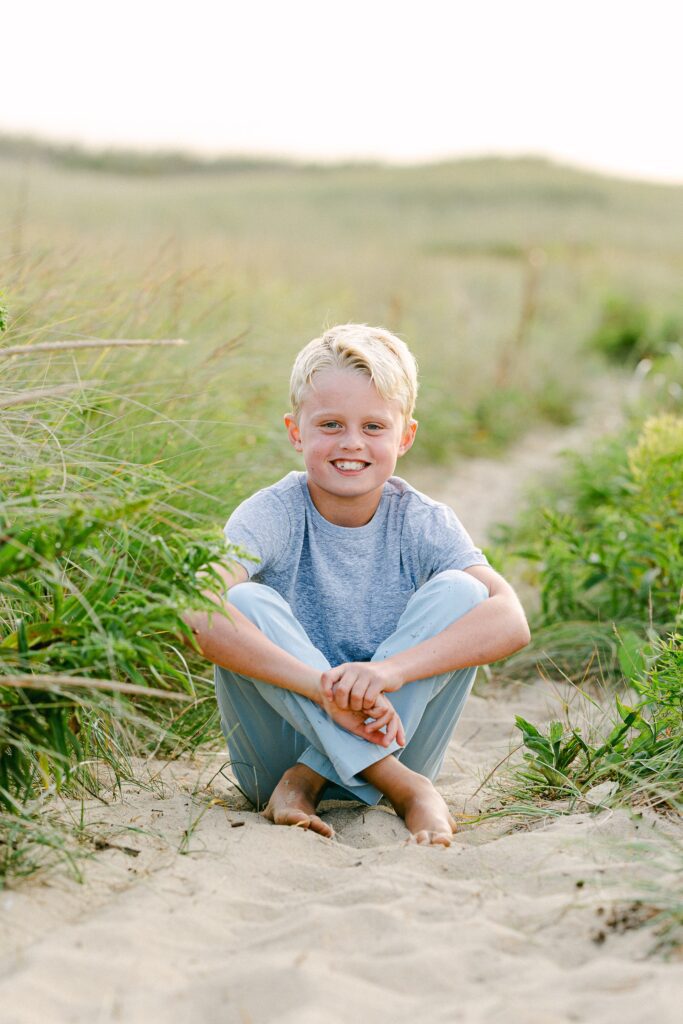 Blond boy sitting cross-legging in the sand, hugging his knees and smiling at the camera. 