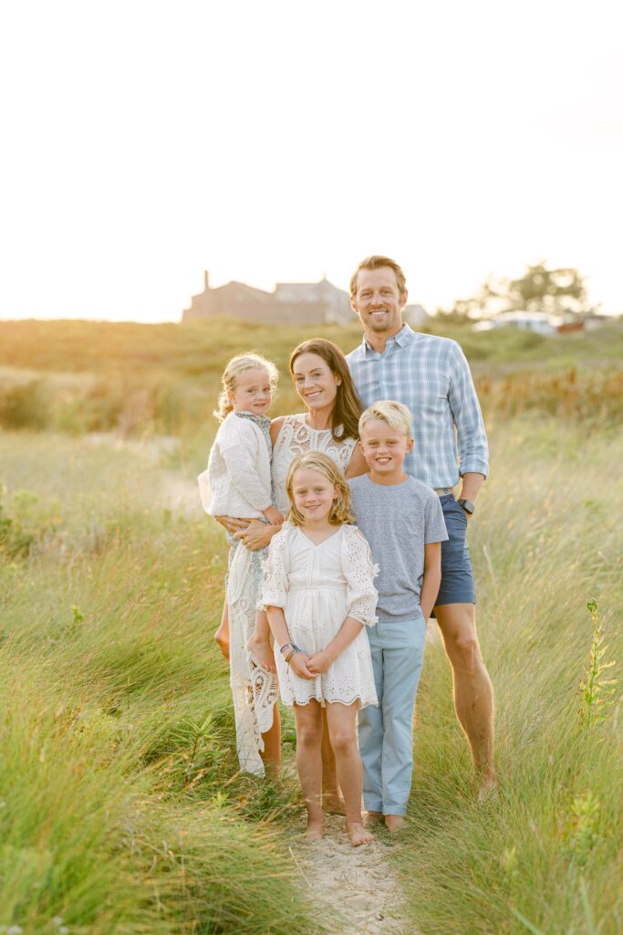 Family of five standing barefoot in the grassy dunes and smiling at the camera. 
