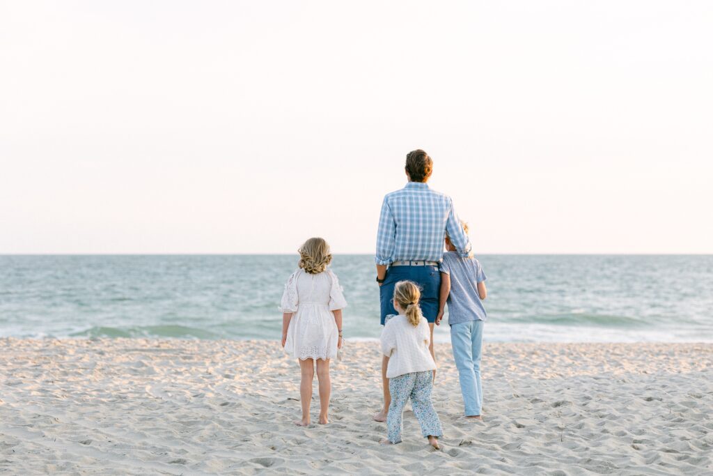 Shot of dad and kids from behind - they are looking at the ocean, dad's arm is around his son, his middle daughter is standing next to him, and his youngest daughter is walking up to them from behind. 