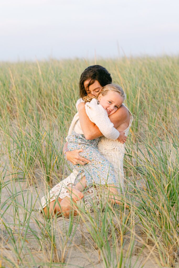Mom kneeling in the sand dunes holding youngest blonde daughter tightly to her and nuzzling her nose into her neck. 