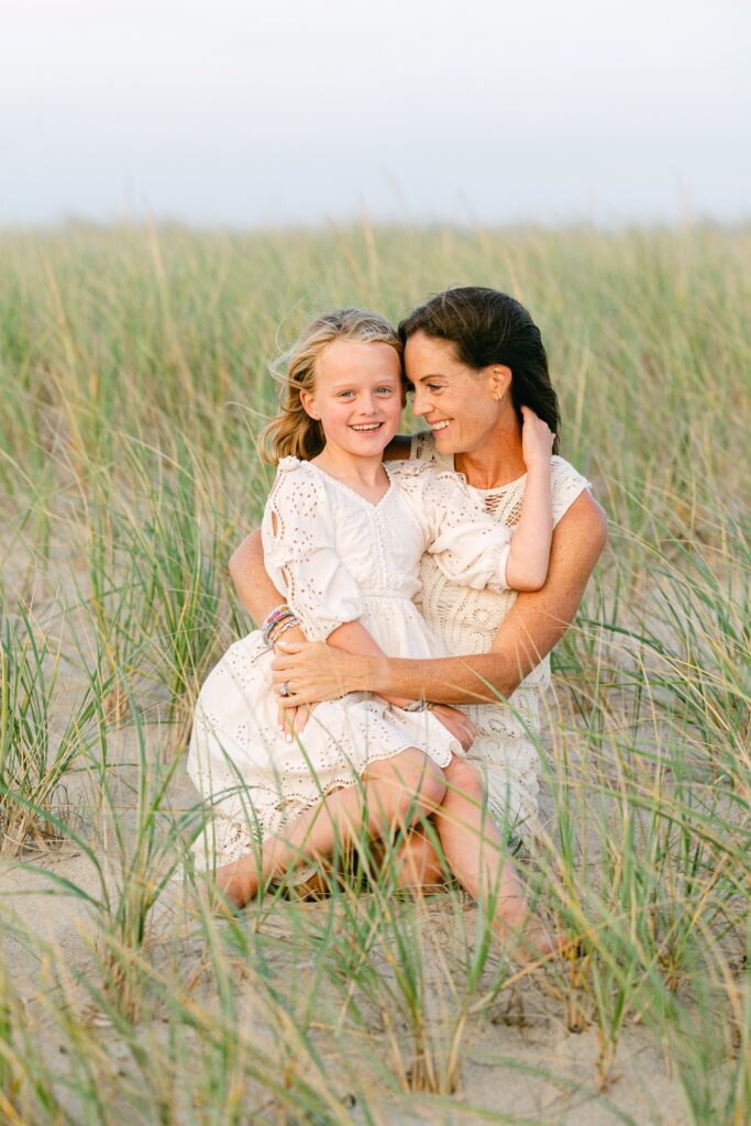 Mom sitting in the sand dunes with arms around daughter who is smiling at the camera, sitting on mom's lap and holding onto mom's neck. 