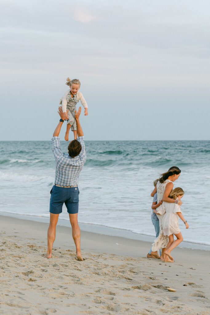 Dad in checkered shirt and blue shorts throws his daughter up in the air on the edge of the ocean on Nantucket, while his wife and older two children play on the beach.