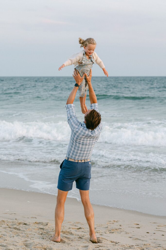 Dad in checkered shirt and blue shorts throws his daughter up in the air on the edge of the ocean on Nantucket.